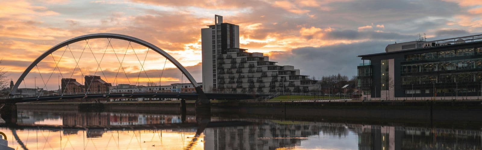 A bridge and the Glasgow skyline sit next to a river that is reflecting a sunset. 